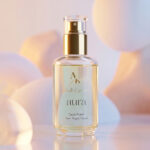 Aura perfume bottle with gold spray top on a reflective surface and a soft pastel background.