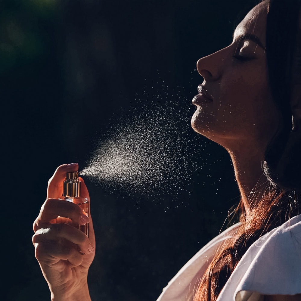 Woman holding a perfume bottle and spraying fragrance, a fine mist dispersing in front of her face against a dark background]