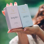 Two pastel perfume boxes labeled 'inspire' and 'aura' held by a person with pink manicured nails outdoors, showing brand logo at top.
