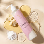 Bottle of Inspire body milk with pump, pink label, surrounded by creamy soaps and powders on a beige background, product shot