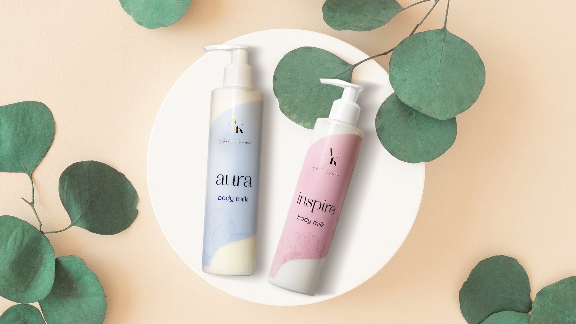 Two pastel-colored body milk bottles on a white plate with green eucalyptus leaves, soft peach background—Aura (blue) and Inspire (pink) labels visible.