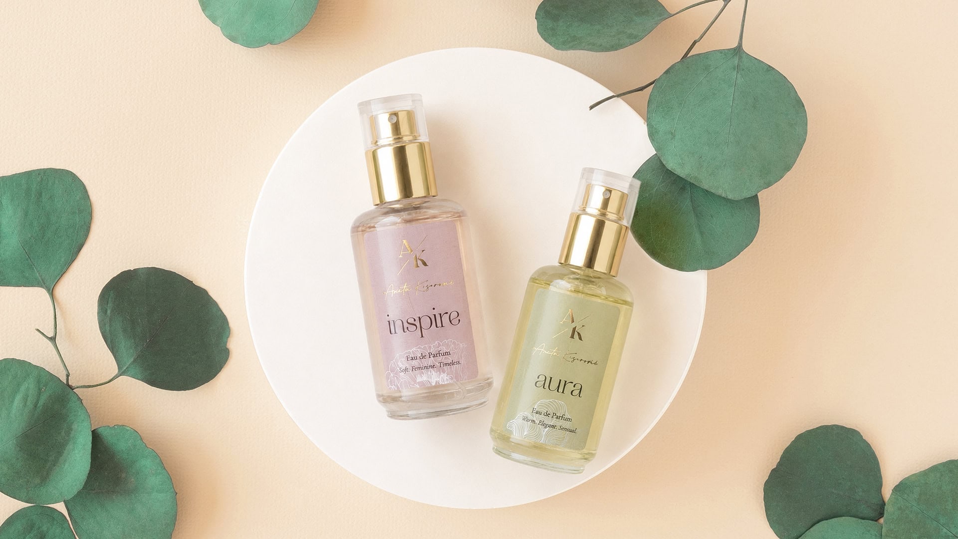 Two perfume bottles with gold caps on a white circular dish, surrounded by eucalyptus leaves against a beige background.