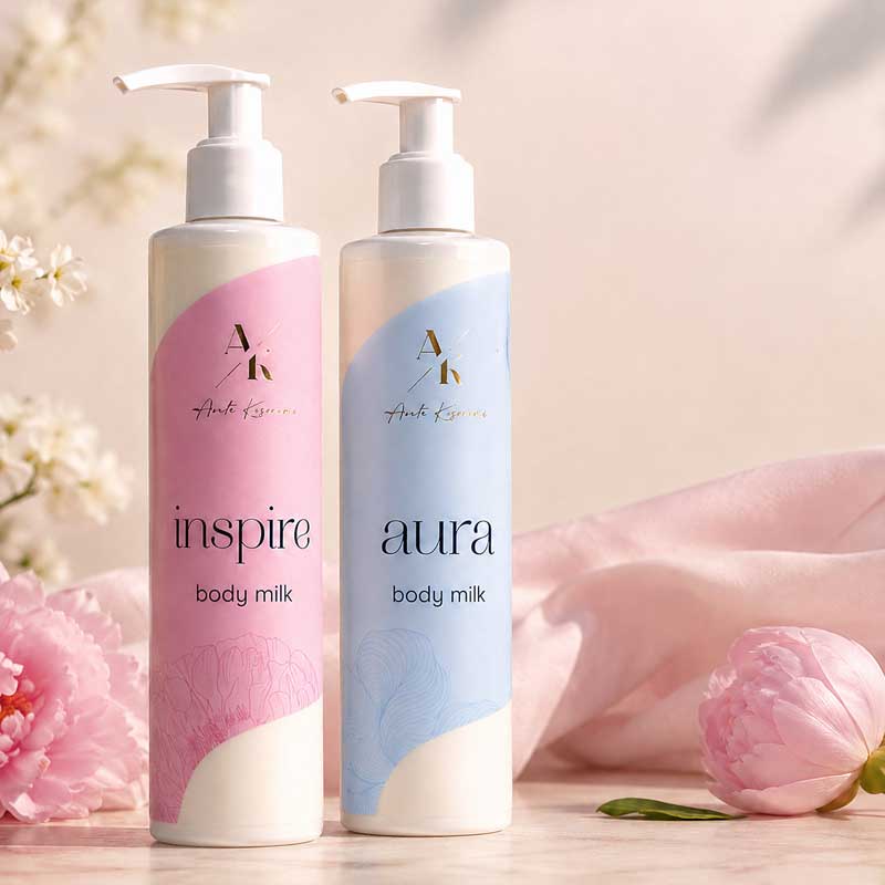 Two pastel-colored pump bottles of Inspire Aura body milk (pink 'inspire' and blue 'aura') on a soft pink background with flowers nearby.