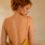 Back view of a woman in a yellow, low-back dress standing in a sunlit bathroom, with a vase of flowers on the counter nearby.