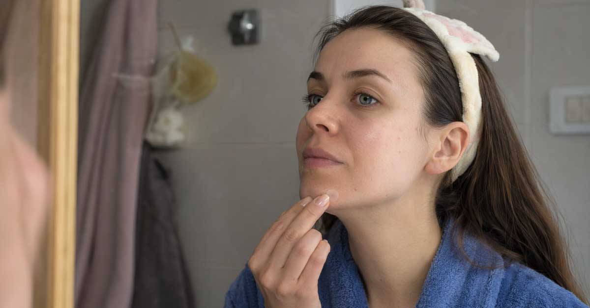 Woman in a blue robe applies moisturizer to her chin as she looks in a bathroom mirror wearing a fluffy headband.